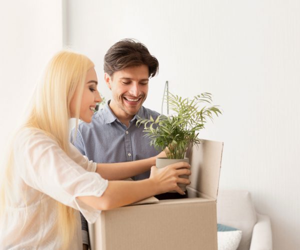 Moving House Concept. Smiling Couple Packing Green Plant In Box. Free Space For Text
